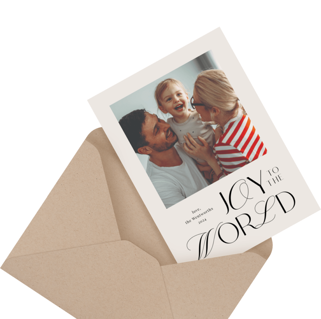 A greeting card with "Joy to the World" features a smiling family of three. The card is partially inserted into a brown envelope.