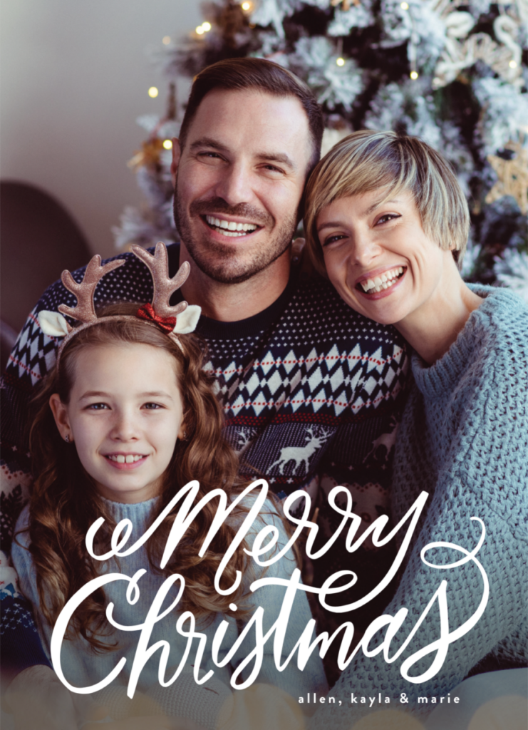 A family of three in festive sweaters smiles in front of a decorated Christmas tree posing for Christmas pictures.