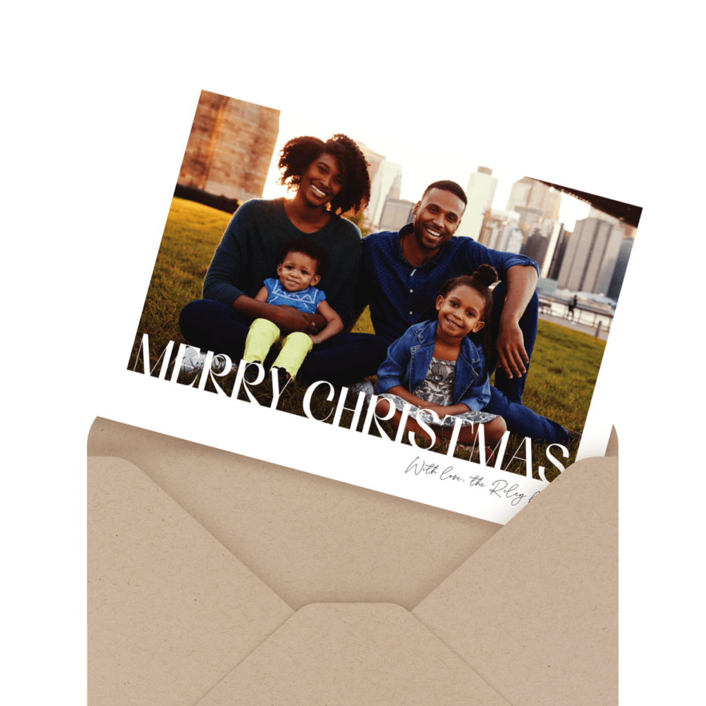 A family of four sitting on grass, smiling. The card says "Merry Christmas" and is placed in an open kraft paper envelope. Buildings and a bridge are in the background.