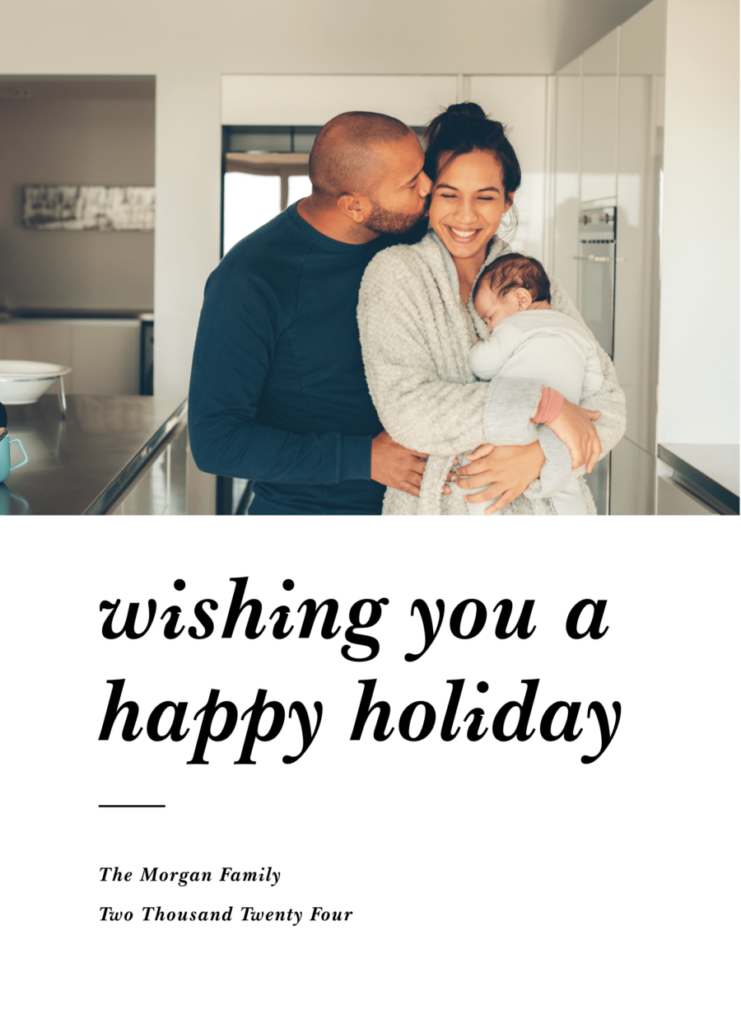 A couple stands in a kitchen, smiling, with the woman holding a baby wrapped in a blanket for their Christmas pictures.