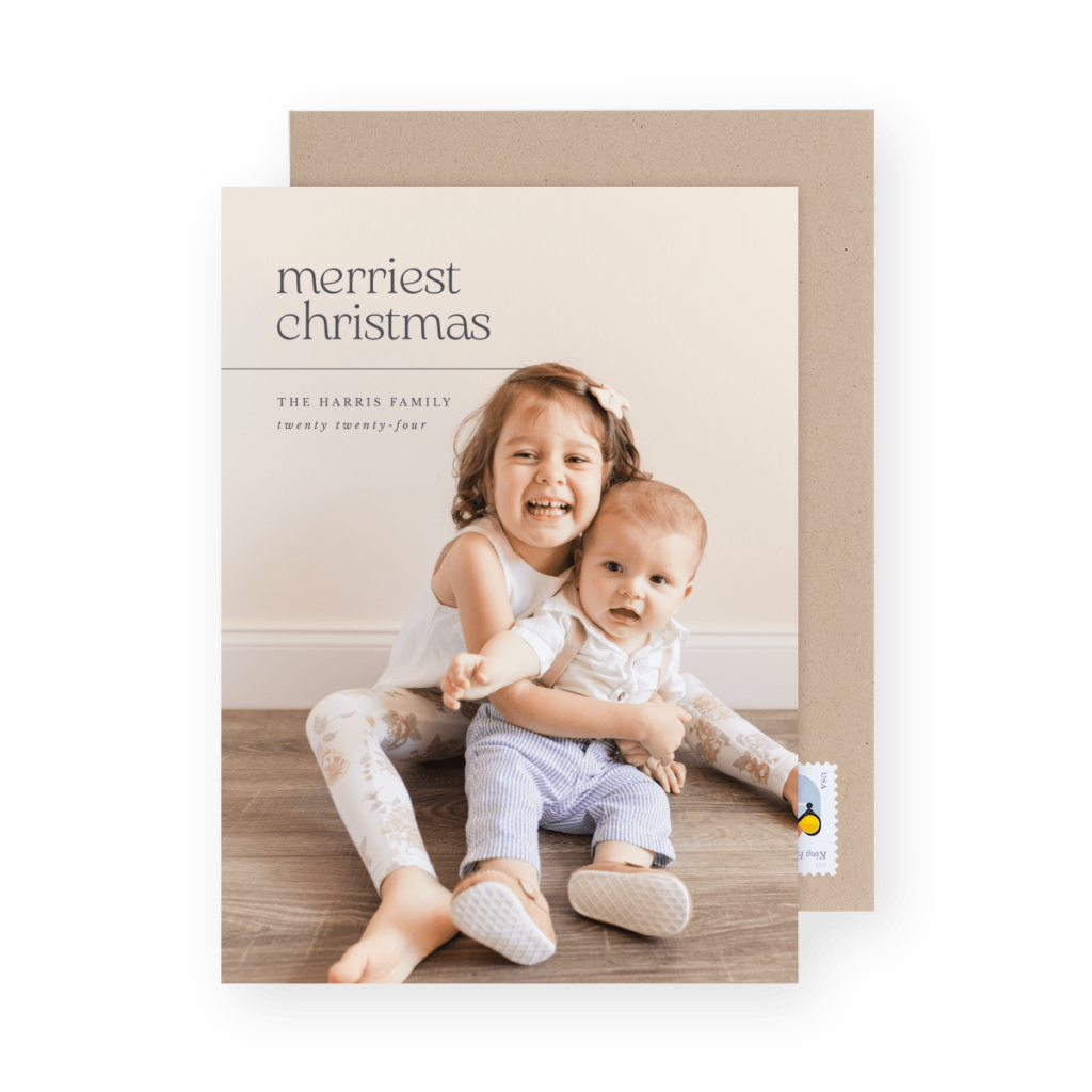 Two young children sit on a wooden floor, smiling and embracing. The card reads "Merriest Christmas."