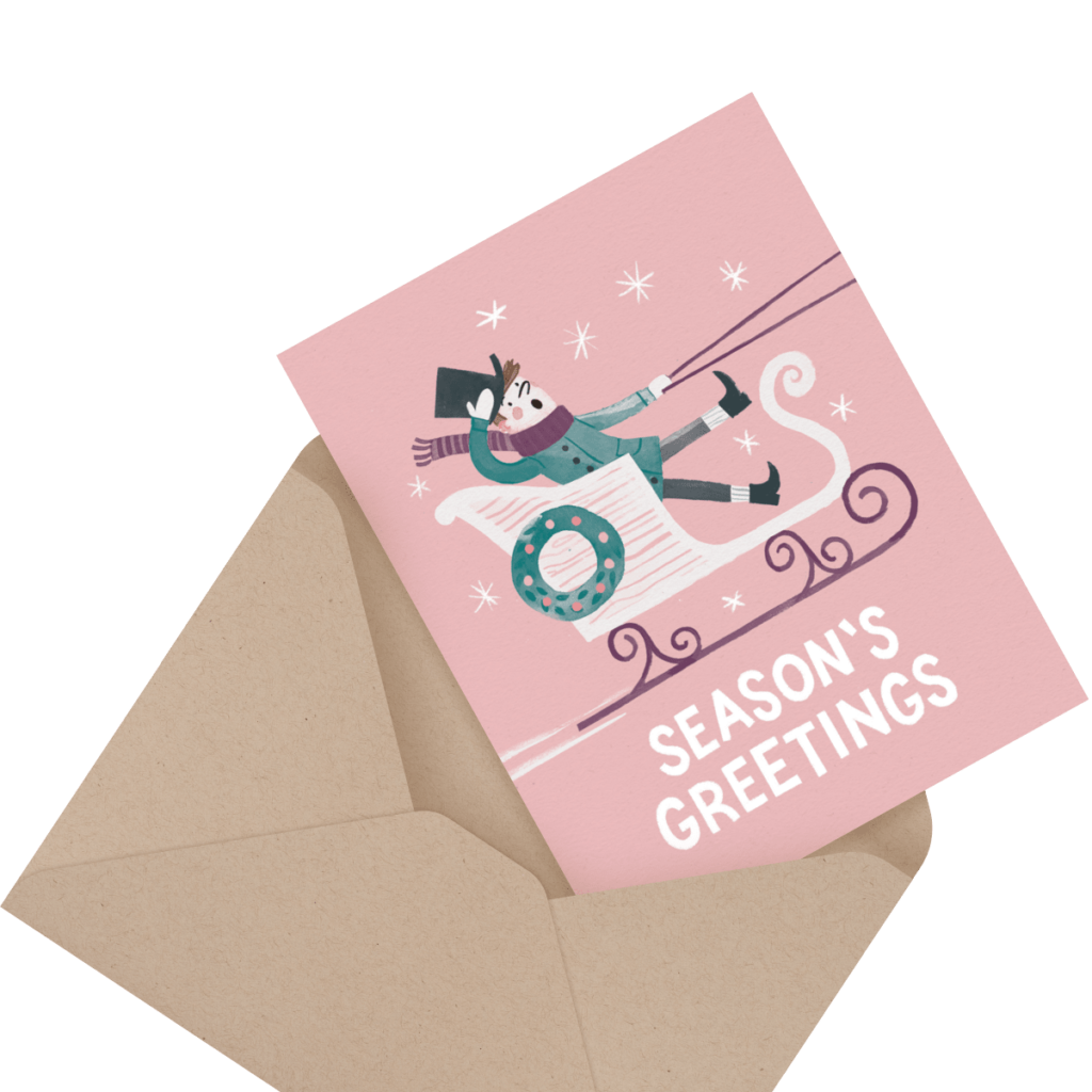 A pink greeting card with "Season’s Greetings" text and an illustration of a person riding a sled, partially inserted in a brown envelope.